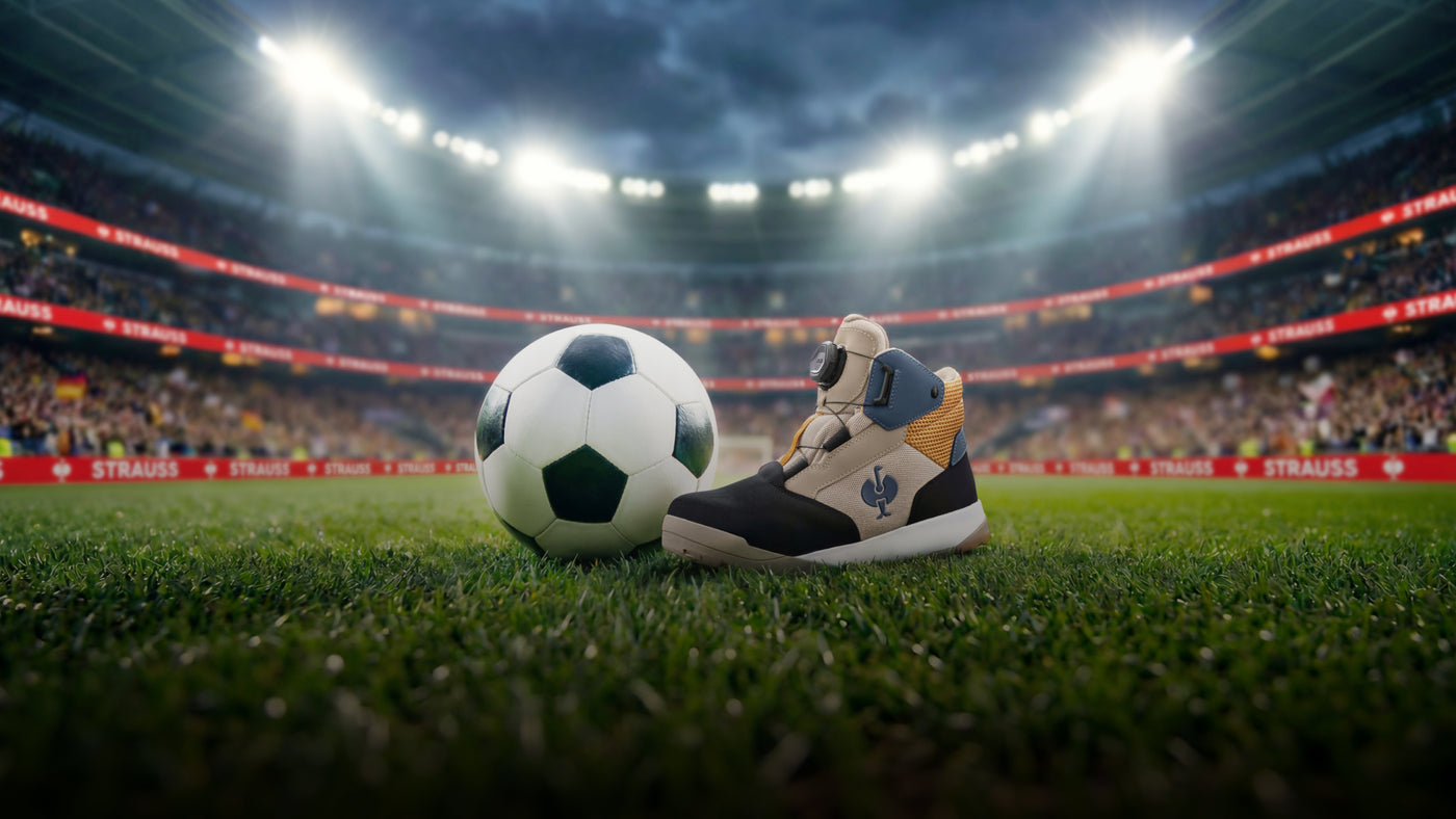 A safety boot in beige, navy, and gray stands on a green stadium field next to a classic black-and-white soccer ball under bright stadium lights.