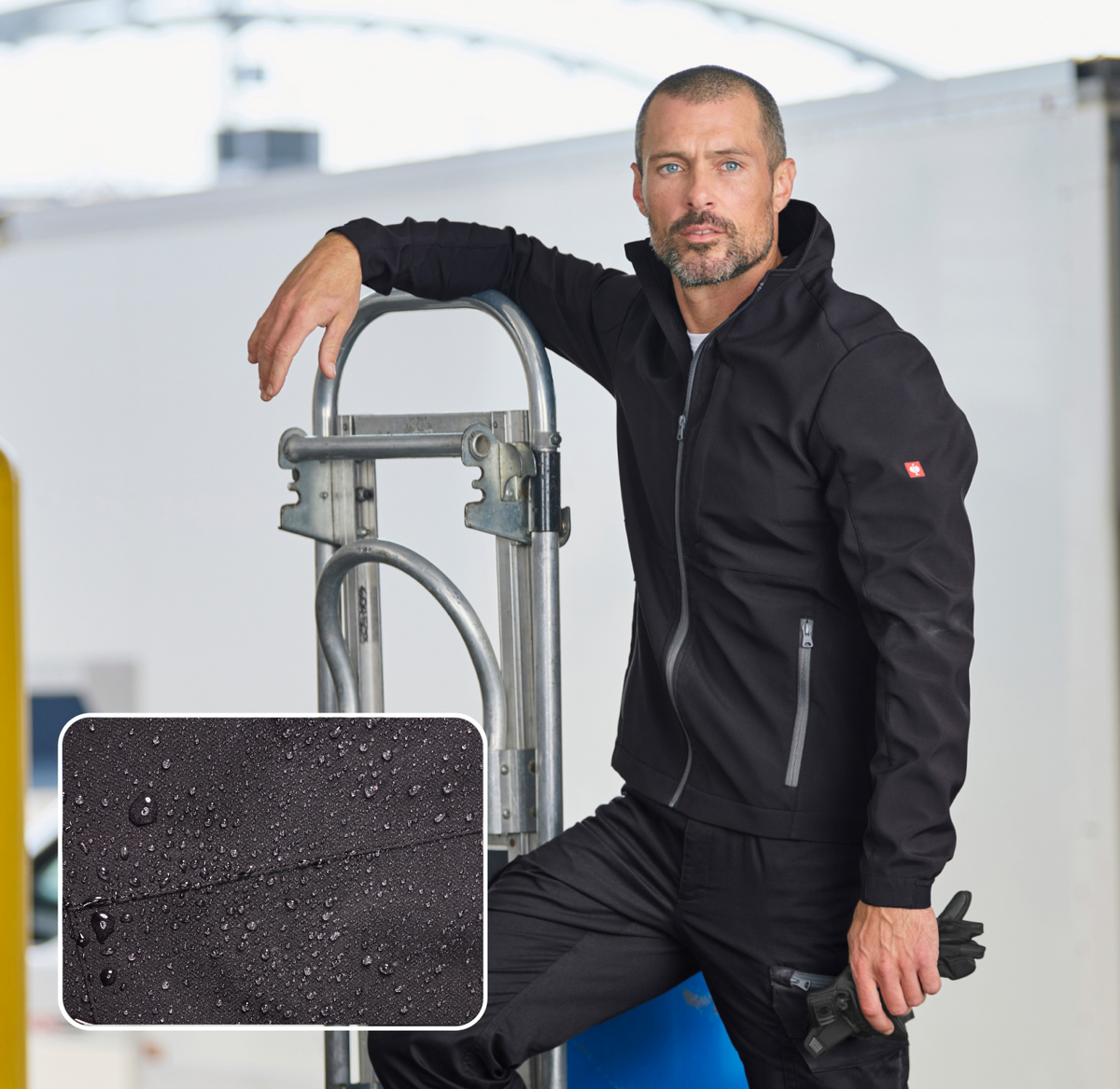 A man in a black zip-up jacket leans on a metal cart in an industrial setting. An inset shows water droplets on black fabric, highlighting the jackets water-resistant material.