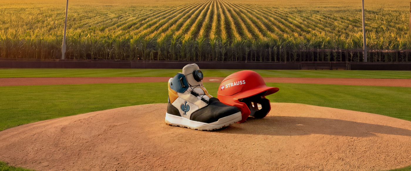 A safety toe work boot in black, white, and blue, paired with a red batting helmet branded 'STRAUSS', resting on a baseball field pitcher's mound with a grassy field and corn fields in the background.