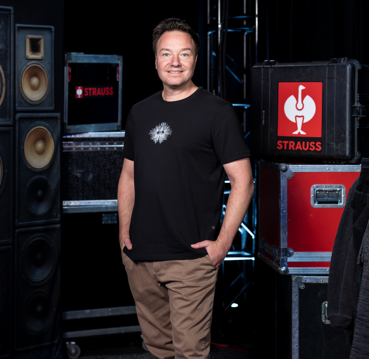 A man in a black t-shirt and beige pants stands smiling with one hand in his pocket. He is indoors, surrounded by large speakers and cases labeled STRAUSS..