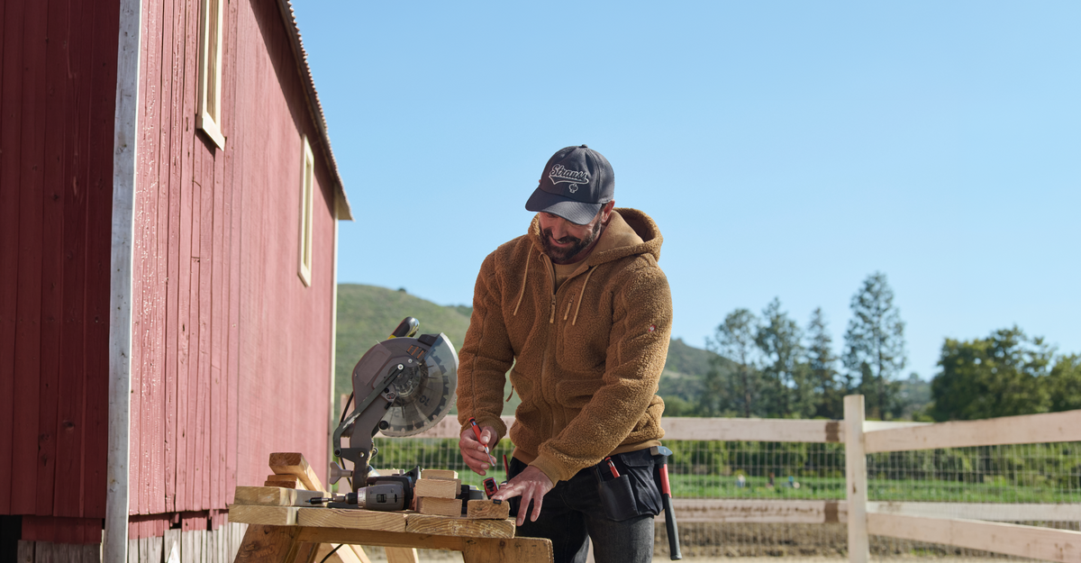 A man wearing a brown sherpa fleece zip-up hoodie, a dark gray and white 'Kings of NY' baseball cap, and black pants is working outdoors next to a red barn, using a miter saw.