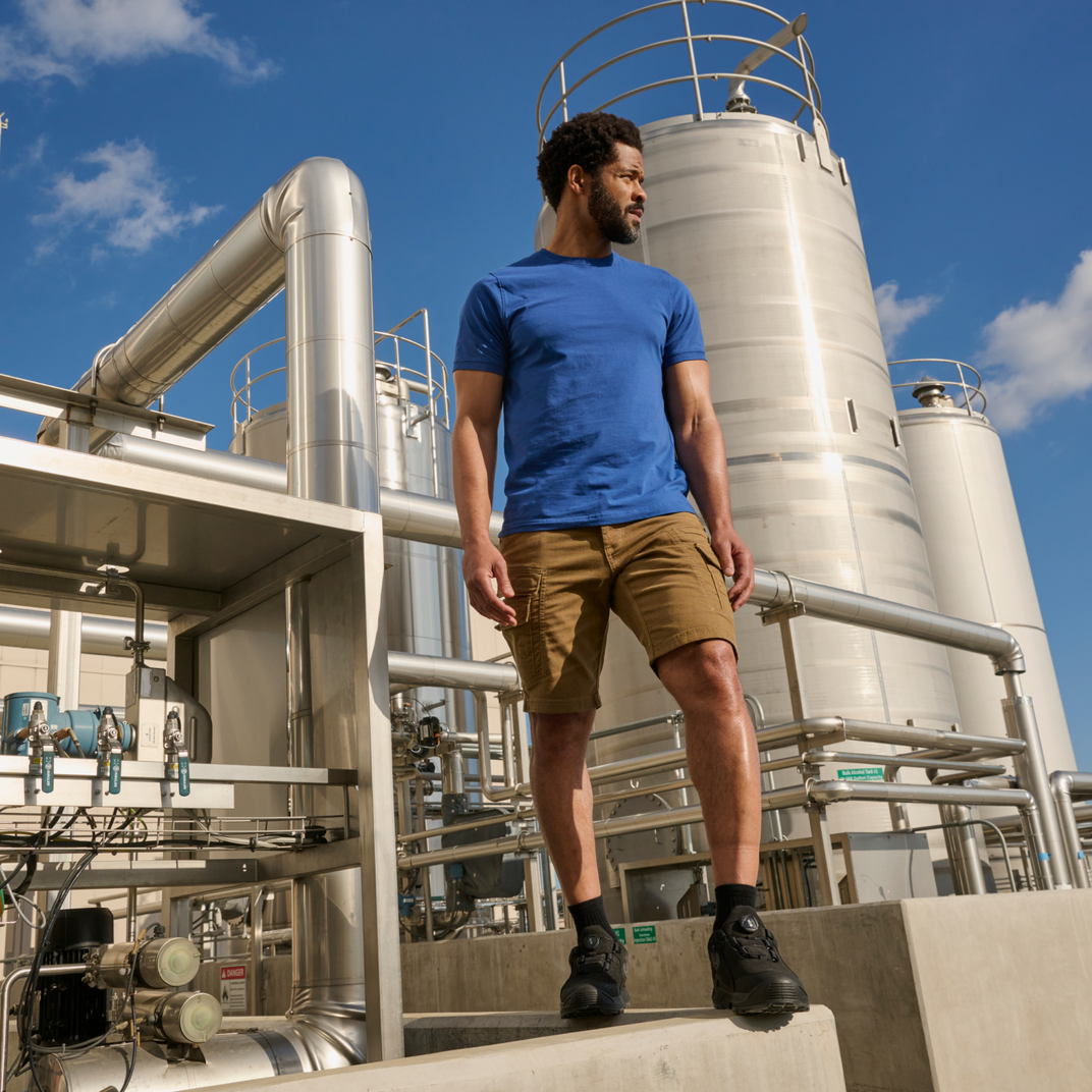 Man in a blue shirt and beige shorts standing on industrial equipment with a clear sky.