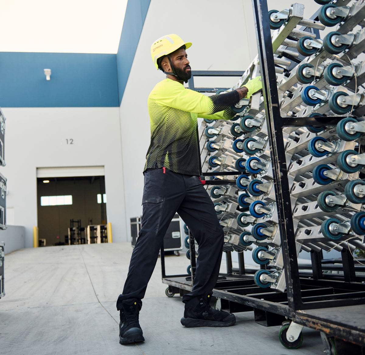 A worker wearing a helmet and safety gear moves a metal rack with multiple wheels outside a warehouse building on a sunny day.