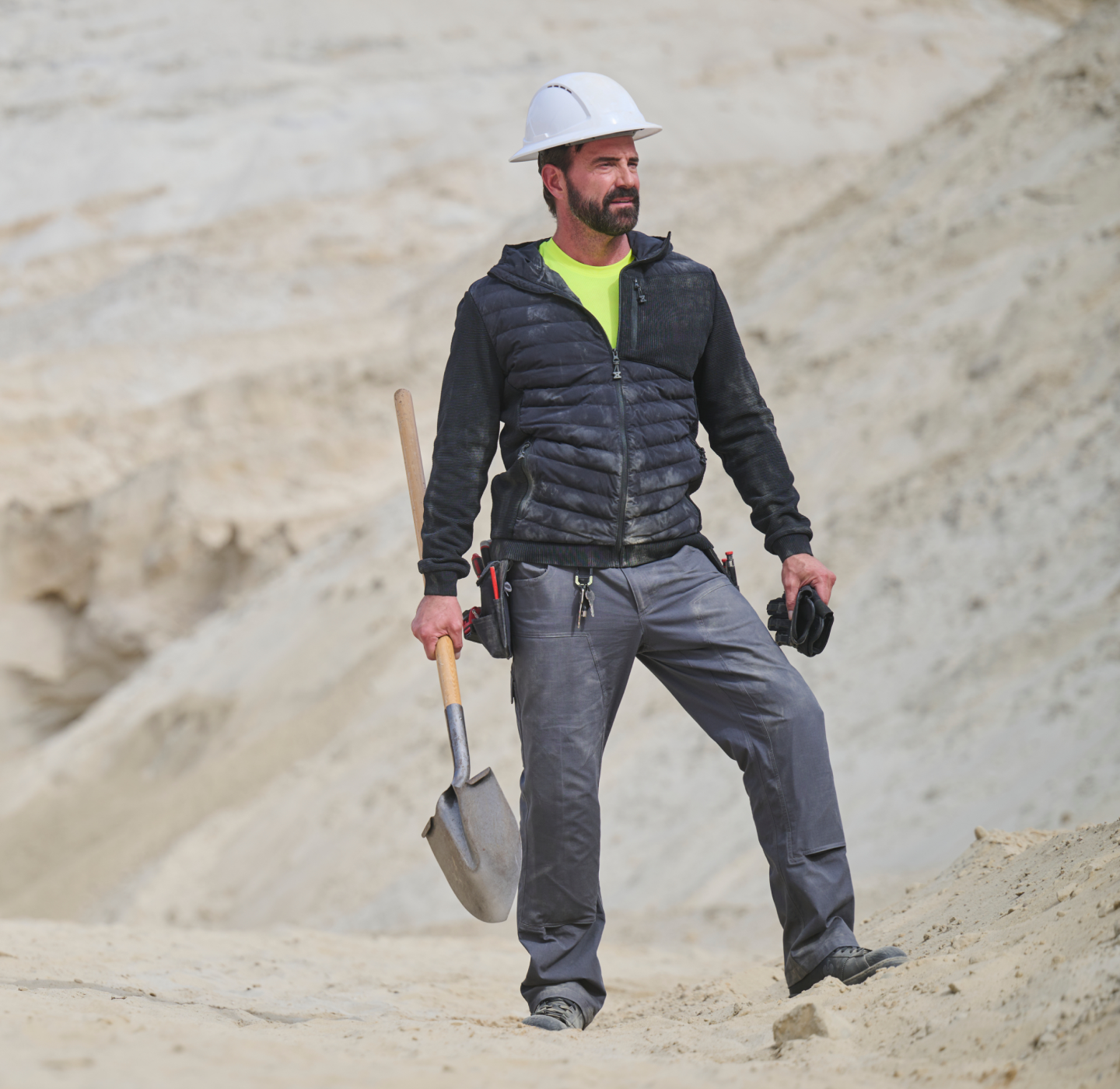 A man wearing a white hard hat and dark jacket stands on sandy ground, holding a shovel. He has a serious expression and carries tools in his pockets, with a rugged construction site background.