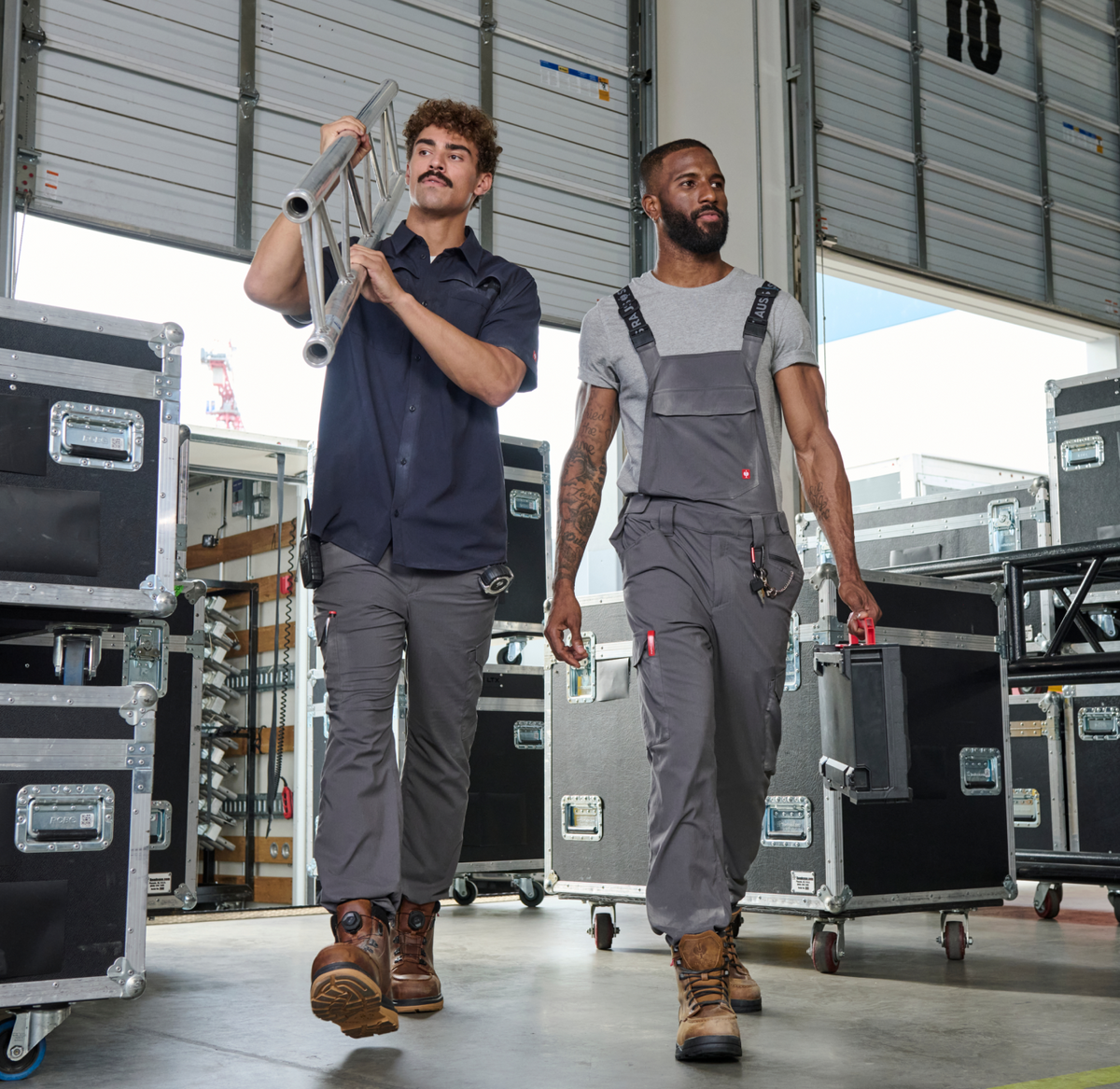Two men in work attire walk through a warehouse filled with equipment cases. One carries a folded ladder, the other holds a toolbox. Large open garage doors and bright natural light are visible in the background.