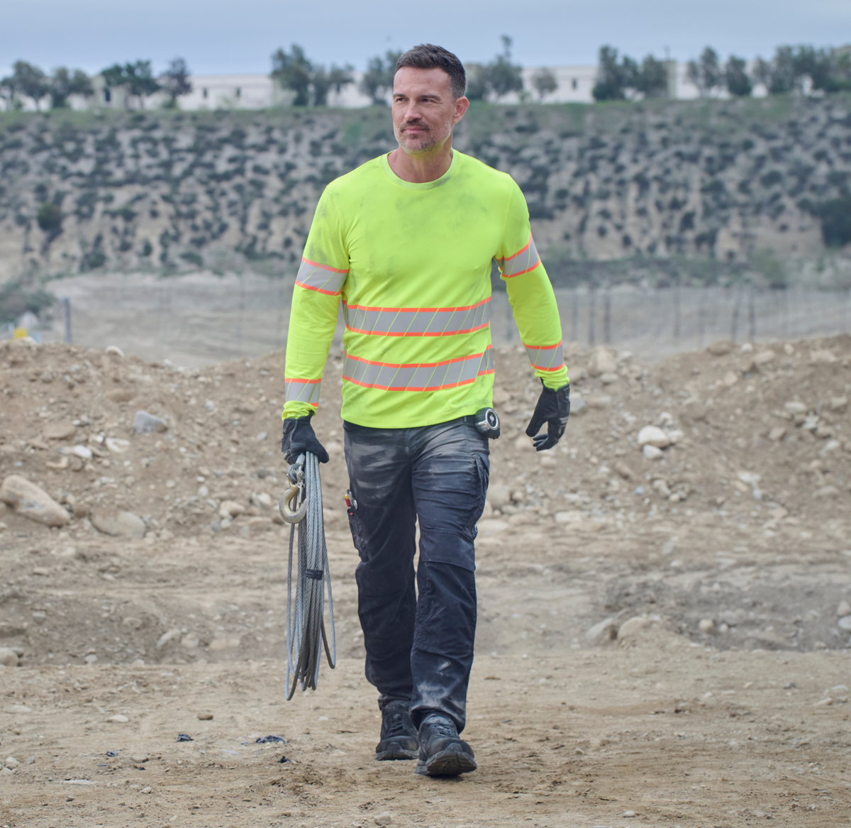 A construction worker wearing a neon yellow reflective shirt and black gloves walks on a dirt site, holding cables in one hand. There are rocky, uneven grounds and a hillside with trees in the background.