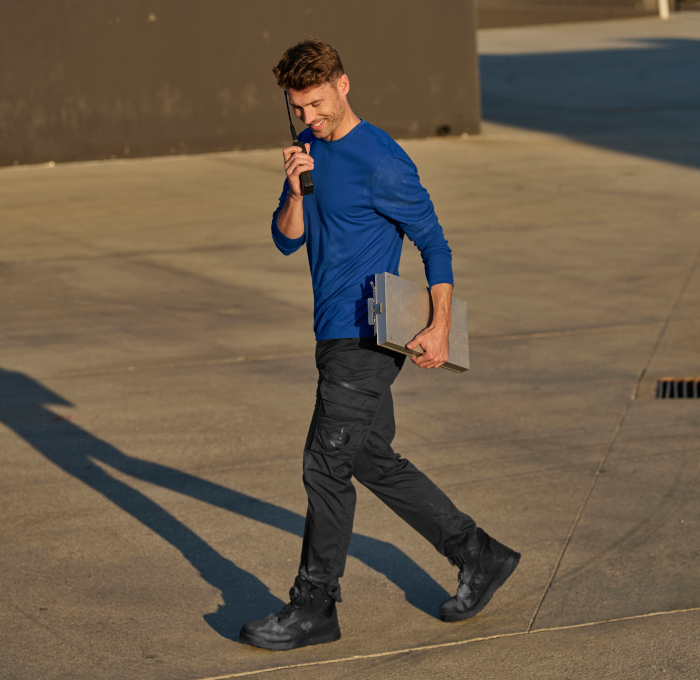 A man in a blue shirt and black pants walks outside on concrete, holding a clipboard and talking on a walkie-talkie, smiling. Shadows and sunlight are visible on the ground.