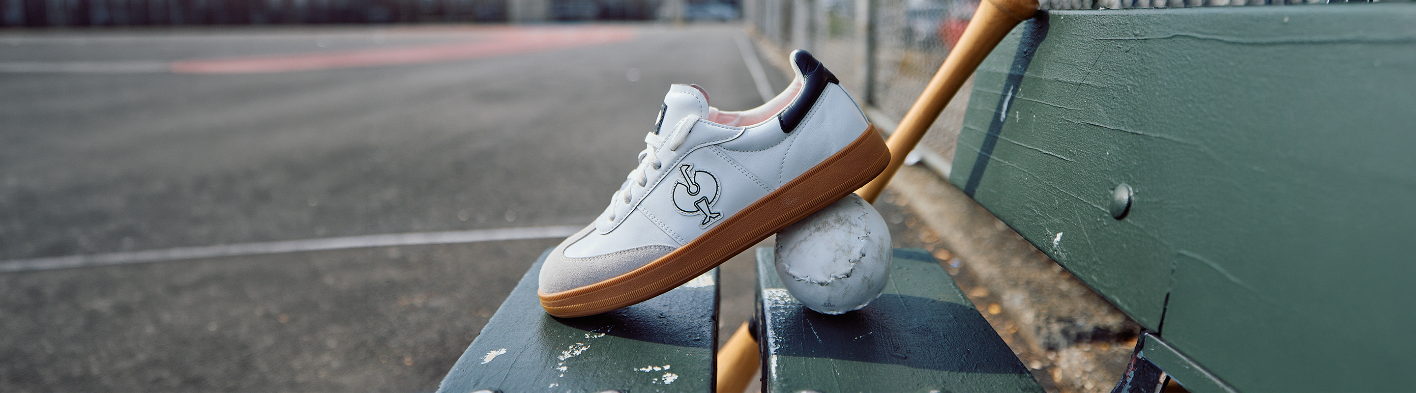 A white sneaker with a brown sole rests on a tennis ball, propped against a green bench on an empty outdoor sports court.