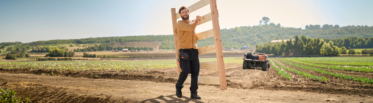 A person in work clothes carries a wooden fence panel along a dirt path near farmland, with green fields and a tractor in the background under a clear sky.