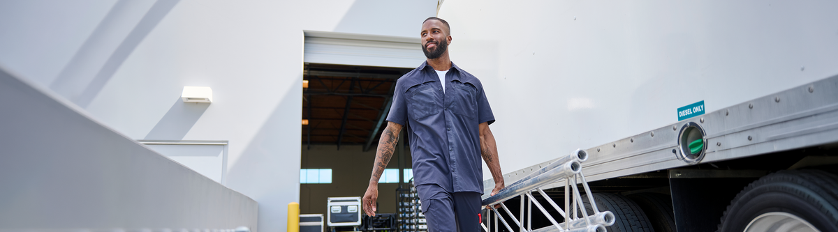 A man in a blue work uniform walks beside a large silver cargo container in a bright industrial setting.