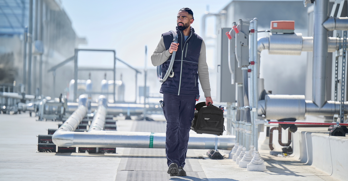 A man walking on a rooftop wearing a navy blue vest, work pants and grey sweatshirt and safety boots.