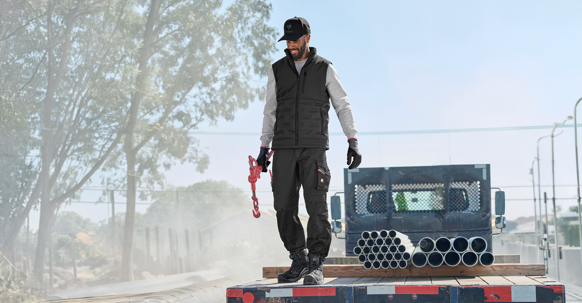 A man wearing a black STRAUSS cap, vest, work pants, gloves and safety boots. He is standing on a truck bed.