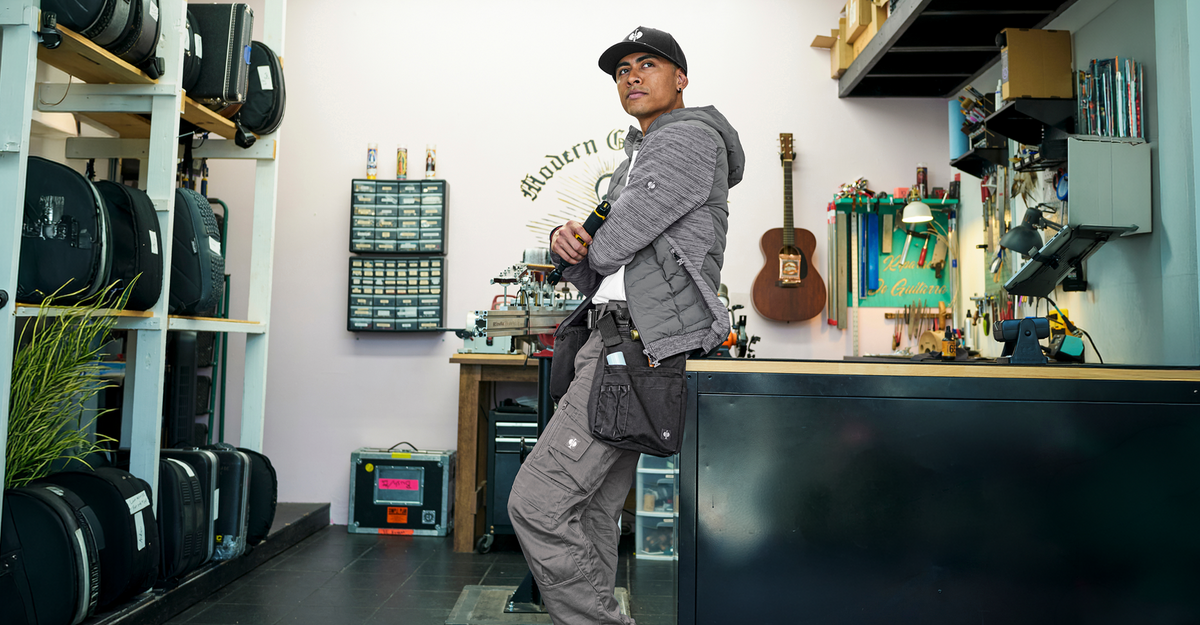 A person in a gray outfit and cap leans against a counter in a workshop with tools, guitars, and equipment visible in the background.