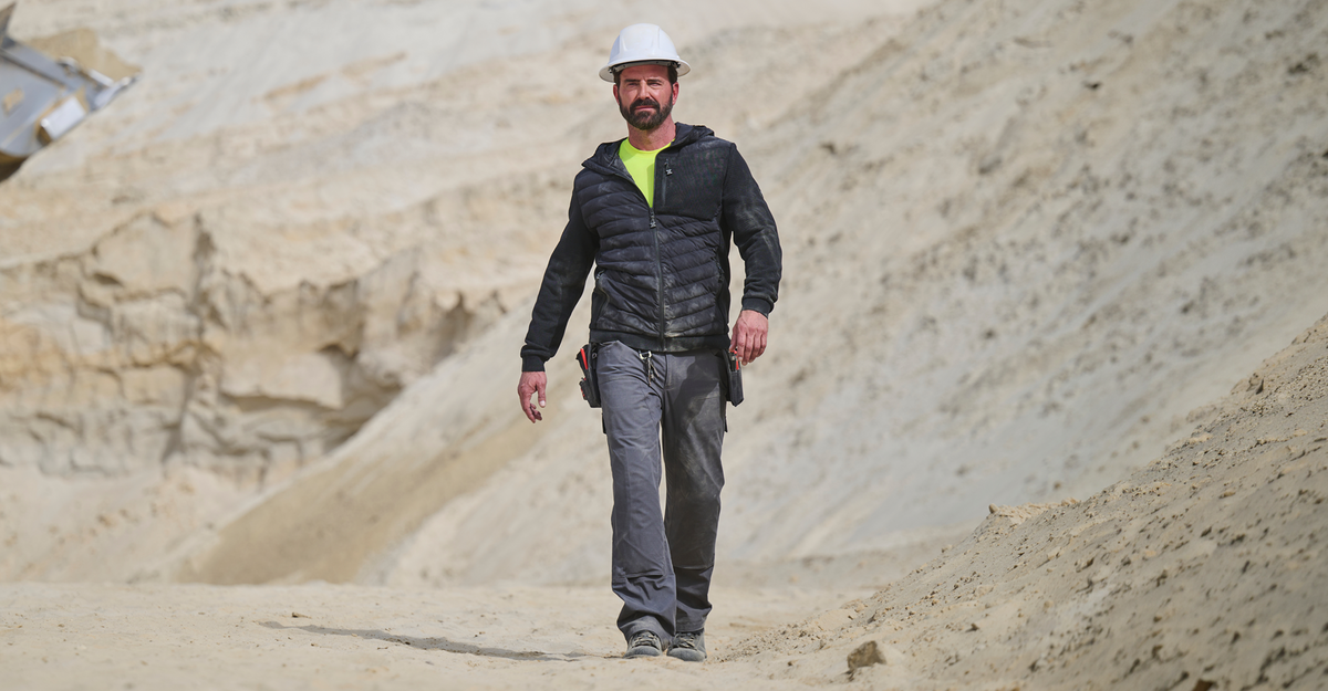 A man wearing a white hard hat, dark jacket, and gray pants walks in a sandy, rocky construction or mining area under daylight.