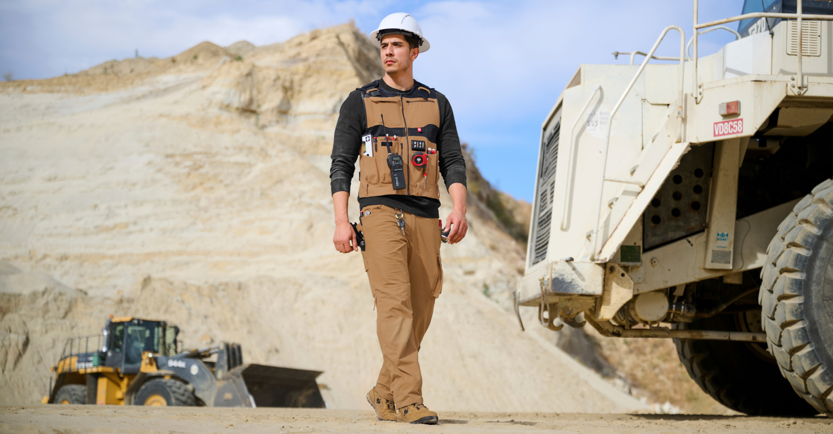 A construction worker wearing a tan utility vest, brown work pants, a black long-sleeve shirt, and a white hard hat walks in a mining site next to a large dump truck.