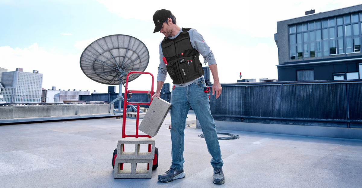 A man wearing a black utility vest and cap stands on a rooftop next to a red step ladder cart, holding a gray box. The background shows a cityscape with a large satellite dish and modern buildings under a bright sky.