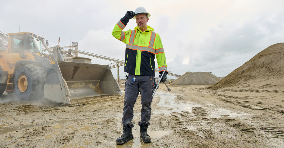 A construction worker wearing a high-visibility neon yellow and navy jacket, gray pants, black boots, and a white hard hat stands in a muddy outdoor site next to a yellow front-end loader under an overcast sky.