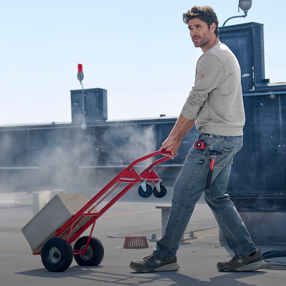 Man pushing a red hand truck on a rooftop with a clear sky.