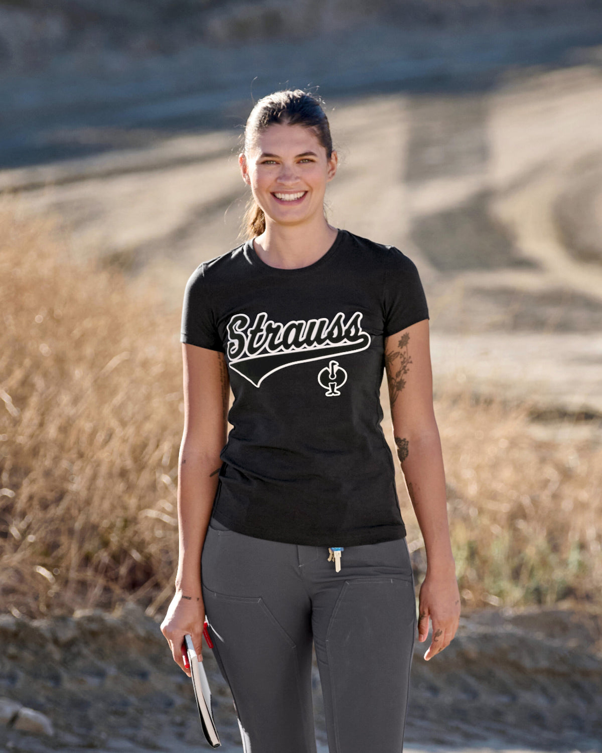 A woman wears the Women's STRAUSS Artwork T-Shirt e.s.e:pic in black with a cap near construction equipment