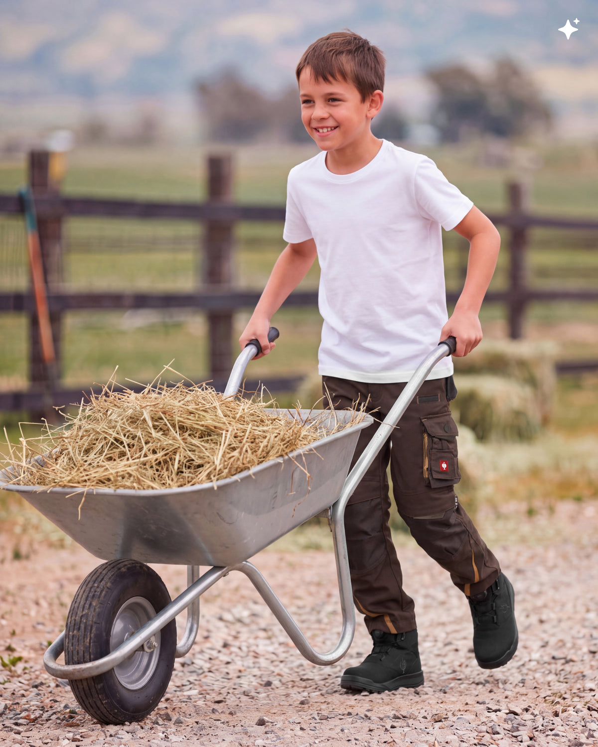 A young boy smiling while pushing a metal wheelbarrow filled with straw on a gravel path in an outdoor farm setting.