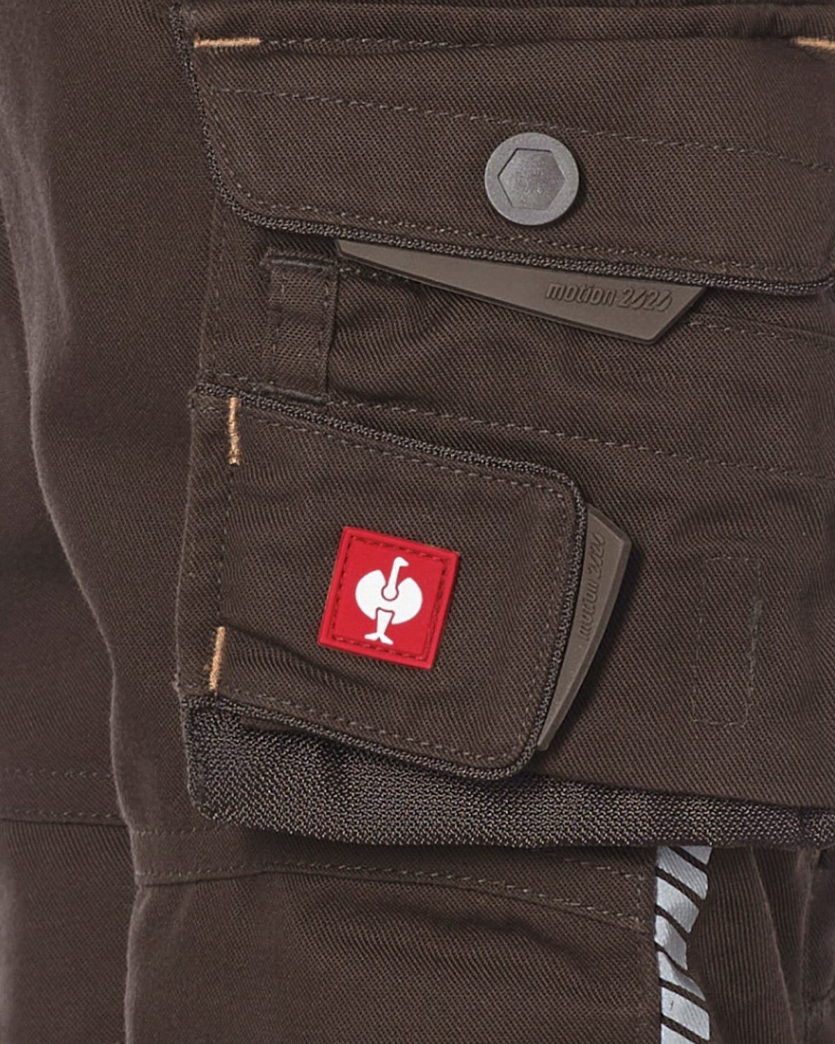 Close-up of dark brown canvas cargo pants showing a front utility pocket with a red square patch featuring a white stylized logo.