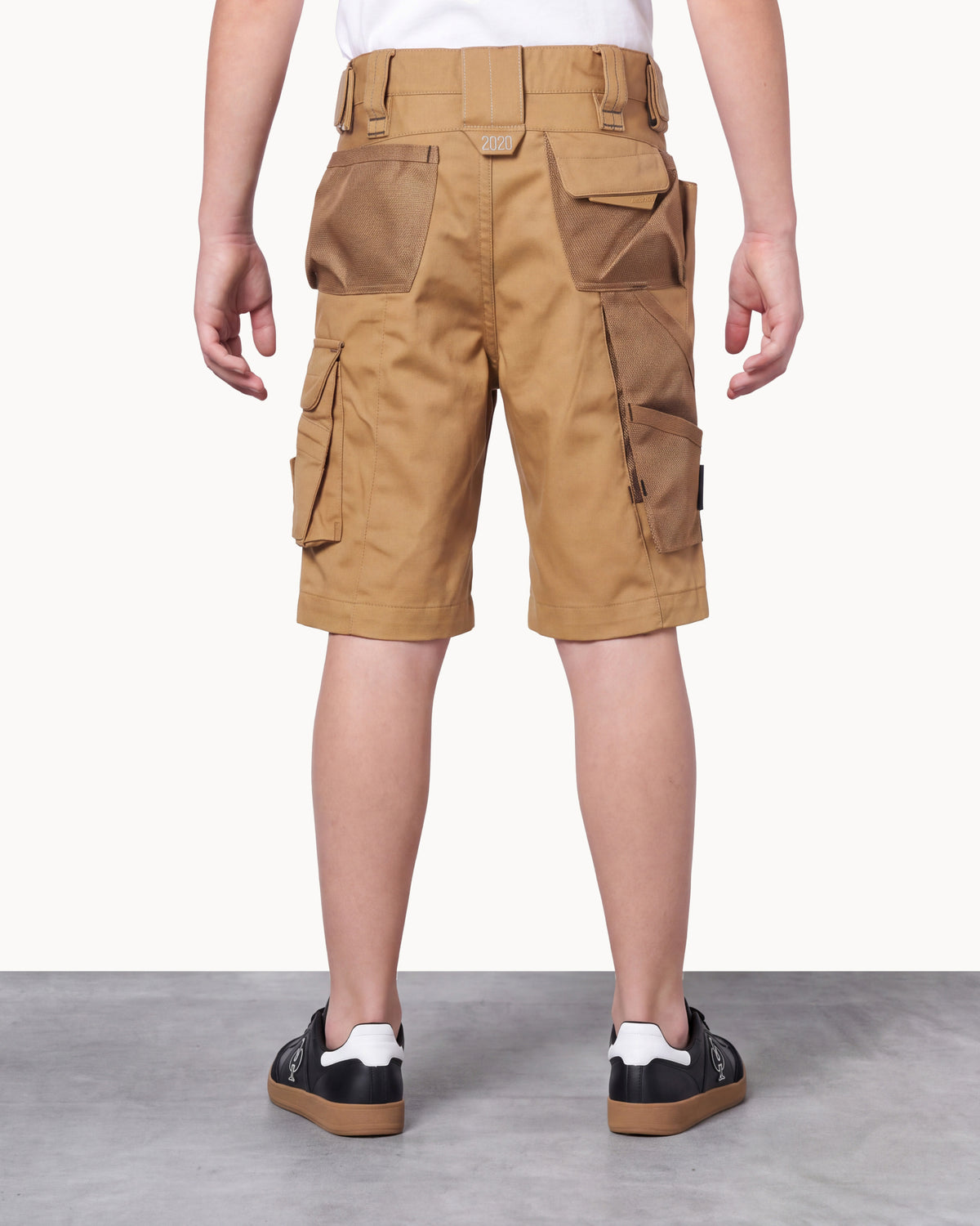 A person shown from the back wearing tan cargo shorts with multiple flap pockets and zippered side pockets, paired with black-and-white sneakers against a plain studio background.