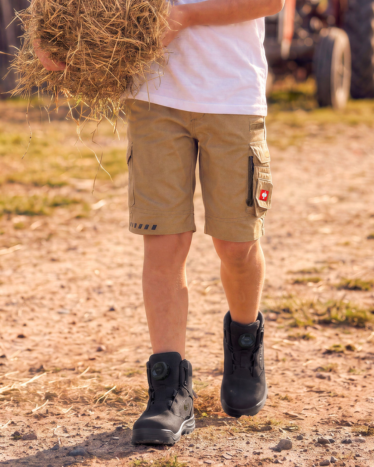 A person outdoors wearing tan cargo shorts, a white t-shirt, and black boots, holding a bale of hay on a dirt path with a tractor blurred in the background.