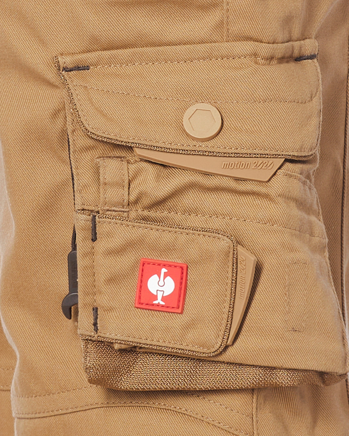 Close-up of a tan canvas utility shorts with a flap pocket featuring a tan snap button and a lower cargo pocket with a red square patch containing a white stylized logo.