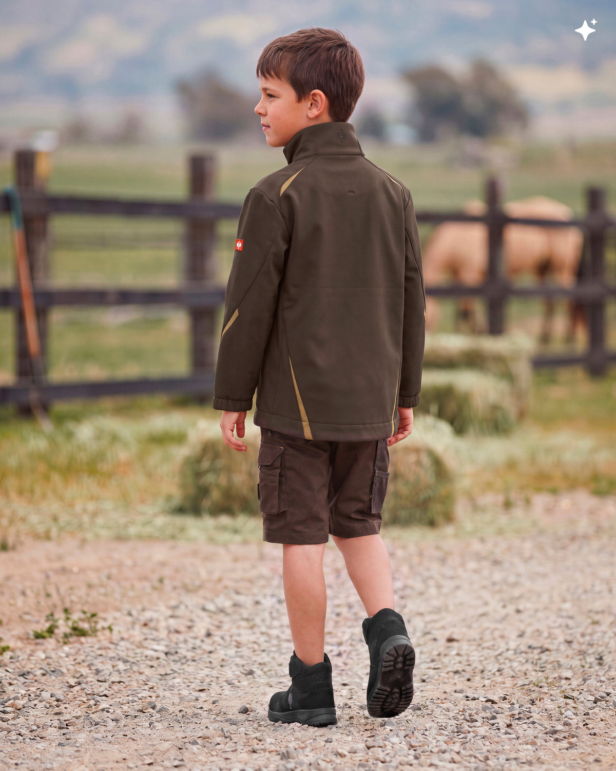 A young boy wearing an olive green outdoor jacket, brown shorts, and black boots stands on a gravel path in a rural setting with a wooden fence and a horse in the background.