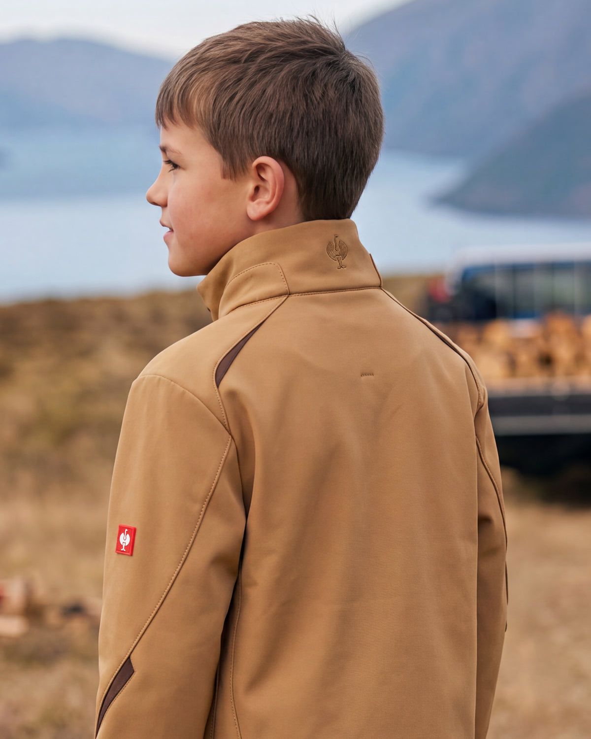 A young boy in profile wearing a tan technical outdoor jacket with dark contrast stitching and a small red logo patch on the sleeve, set against a blurred natural background of water and mountains.