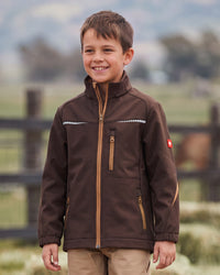 A young boy smiling outdoors wearing a brown softshell jacket with tan zipper details and khaki pants, set against a blurred background of a fence and trees.