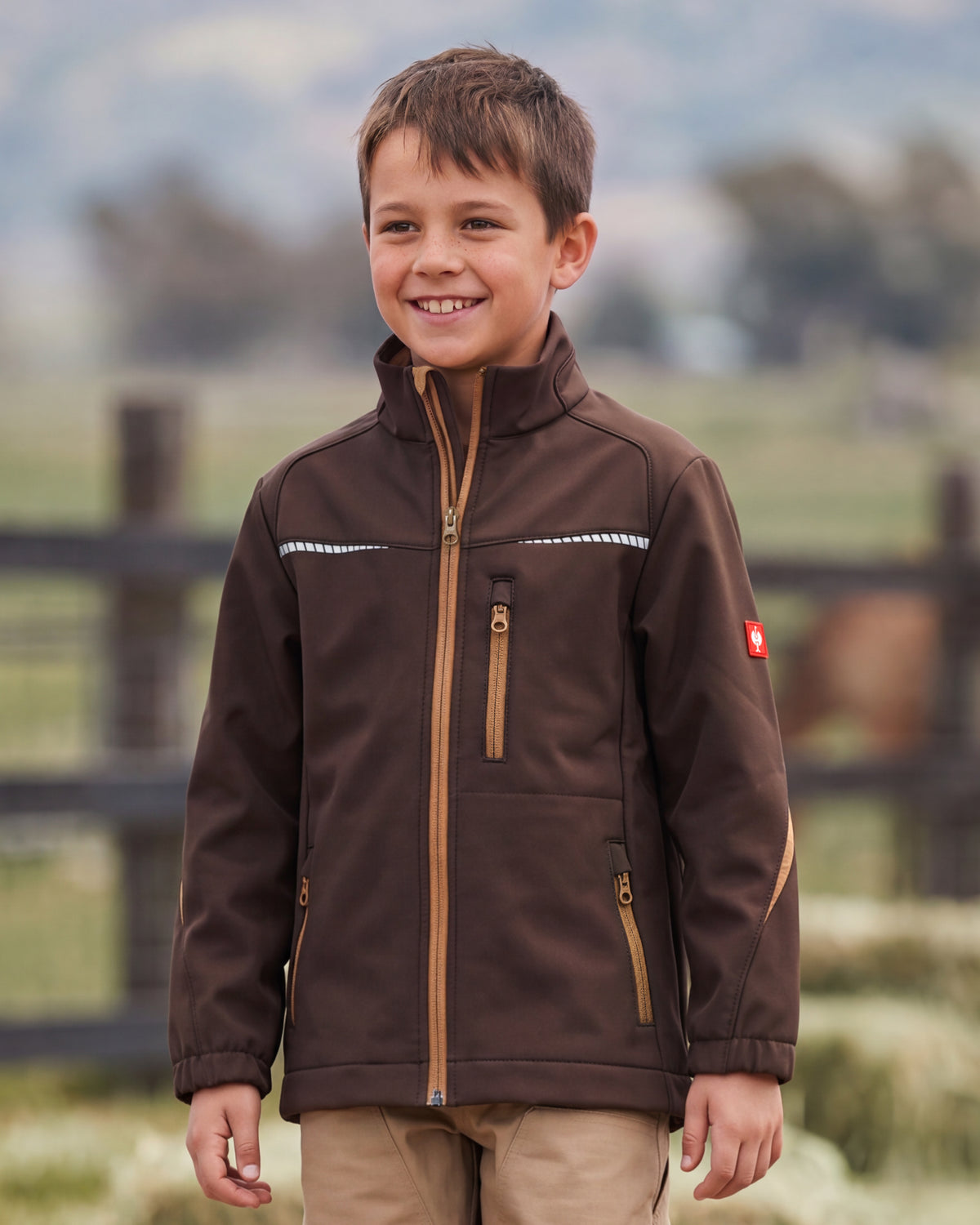 A young boy smiling outdoors wearing a brown softshell jacket with tan zipper details and khaki pants, set against a blurred background of a fence and trees.