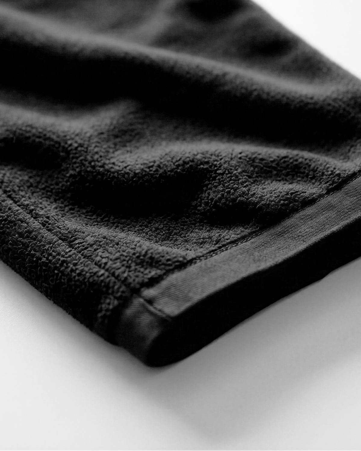 A close-up shot of a black textured fabric, likely a towel or blanket, showing its soft terry cloth texture and stitched edge against a plain white background.