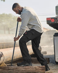 A man in a gray long-sleeve shirt and black work pants bends over while using a long-handled tool near large metal pipes in an outdoor industrial setting.