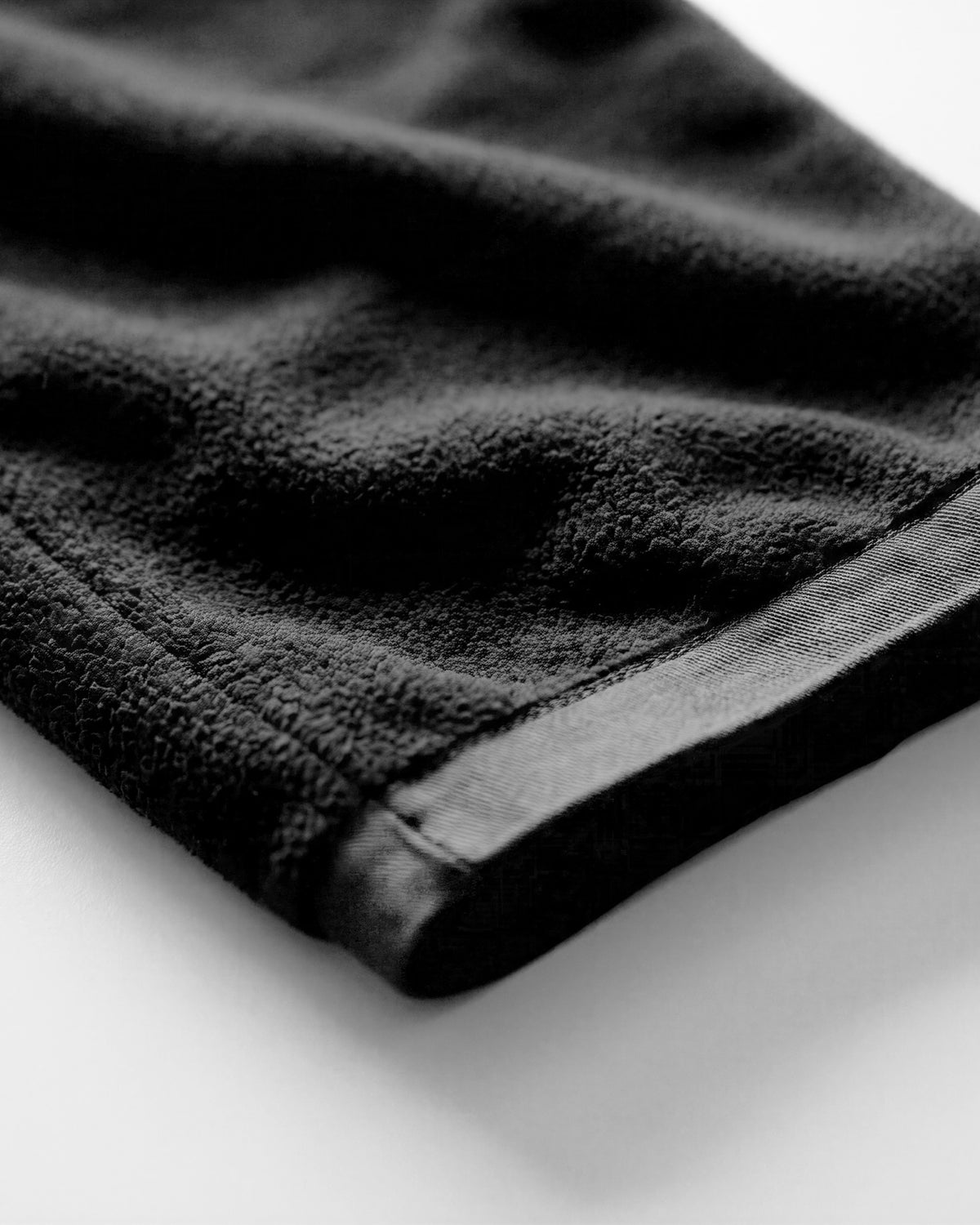 Close-up of a black textured fabric product, likely a towel or blanket, showing the soft terry texture and stitched edge on a light background.