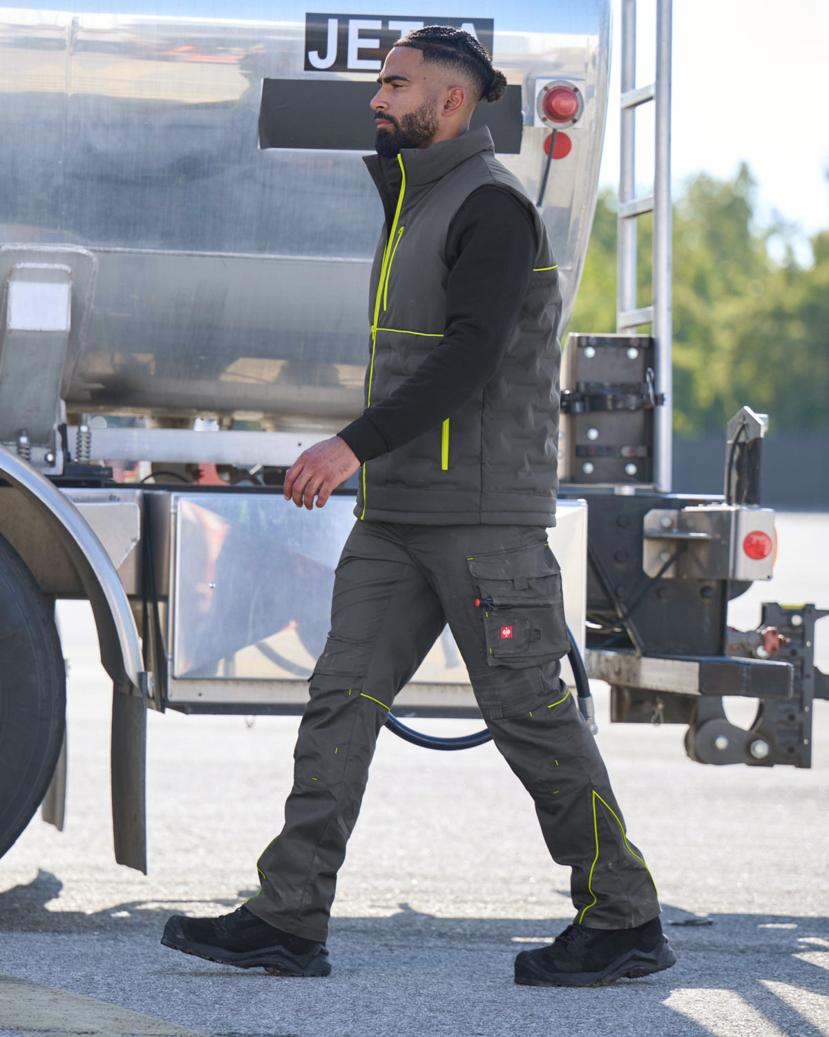 A man wearing a dark gray work vest and matching cargo pants with bright yellow-green reflective trim walks past a large silver tanker truck outdoors.