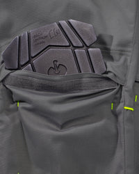 Close-up of a dark gray technical jacket pocket showing a textured black protective pad and bright neon green stitching details.