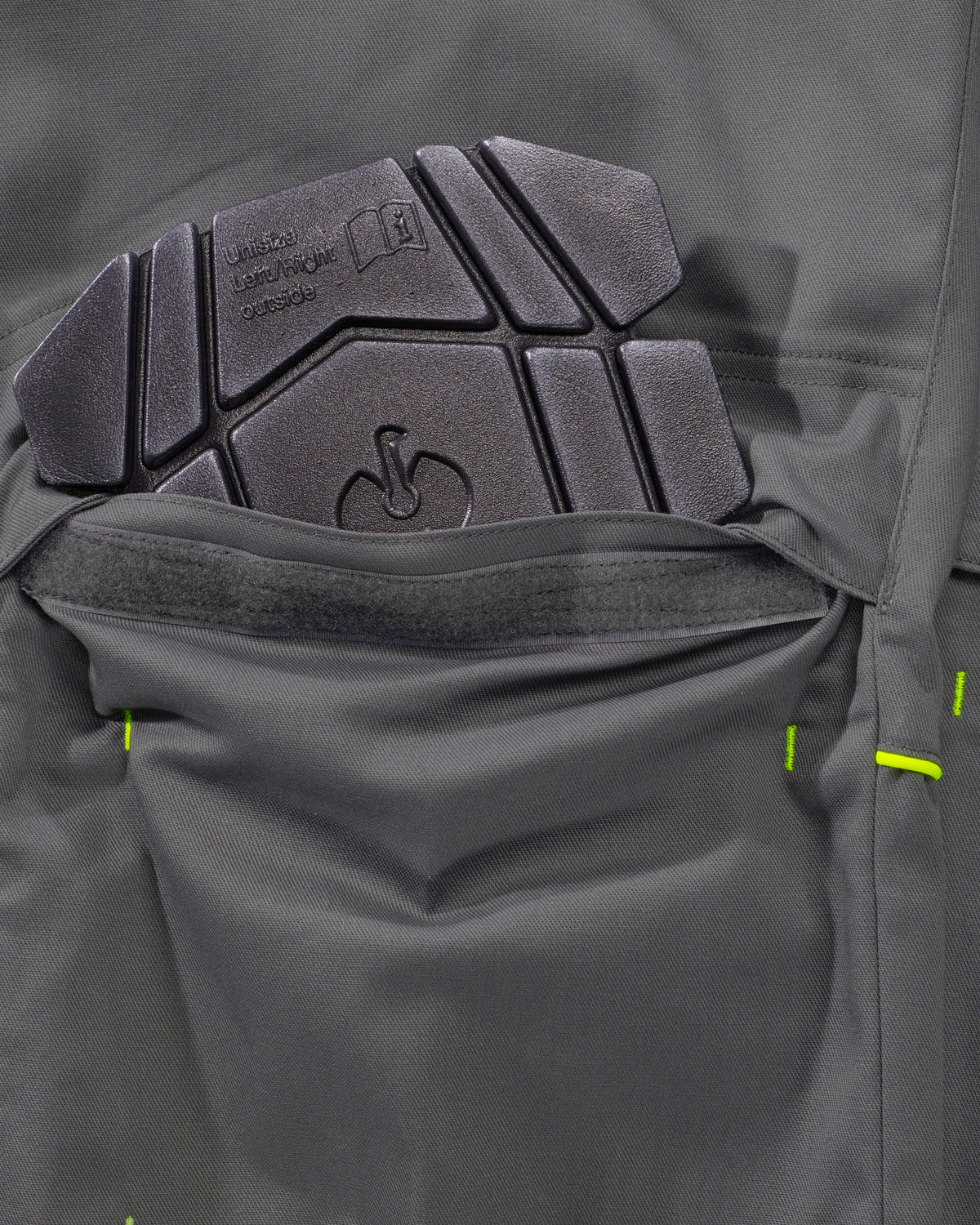Close-up of a dark gray technical jacket pocket showing a textured black protective pad and bright neon green stitching details.