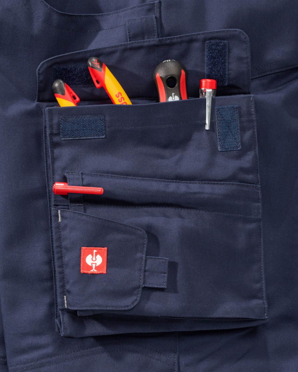Close-up of a navy blue work jacket pocket holding various hand tools, featuring a small red square patch with a white rooster logo on the front flap.