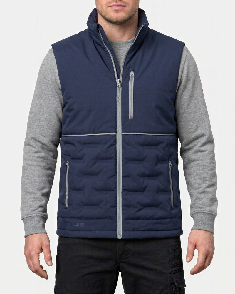 A man wearing a navy blue quilted vest with gray sleeves and gray zipper details, layered over a gray long-sleeve shirt and black pants, against a light gray background.