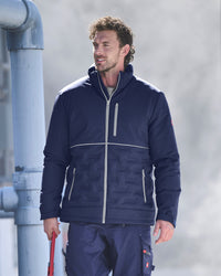 A man wearing a navy blue insulated jacket with light gray zippers and matching navy pants, standing outdoors next to a large gray pipe.