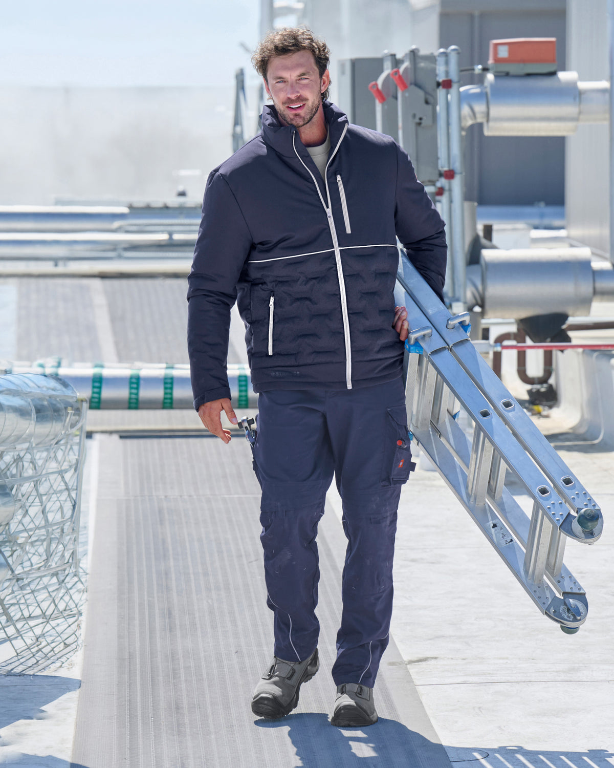 A man wearing a dark navy blue technical jacket and matching work pants walks on a metal walkway while carrying a metal ladder.