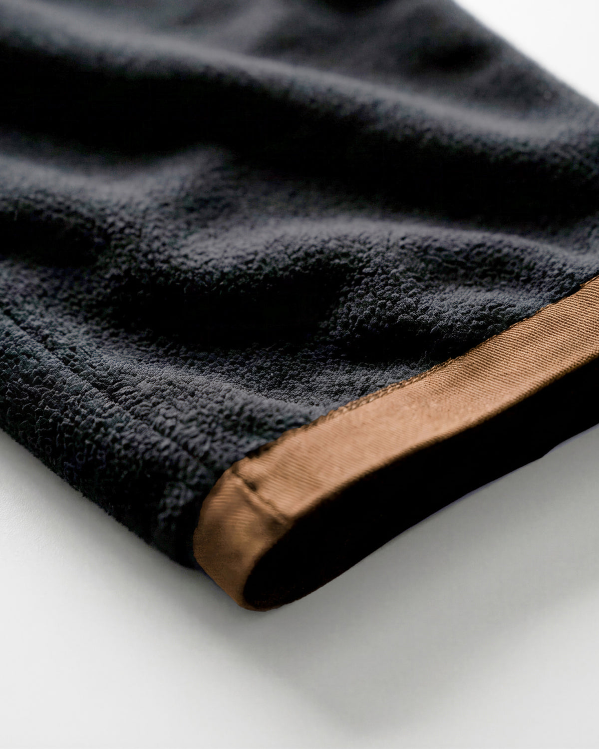Close-up of a dark charcoal gray fleece or terry cloth fabric with a contrasting tan-brown stitched trim, shown on a light gray background.