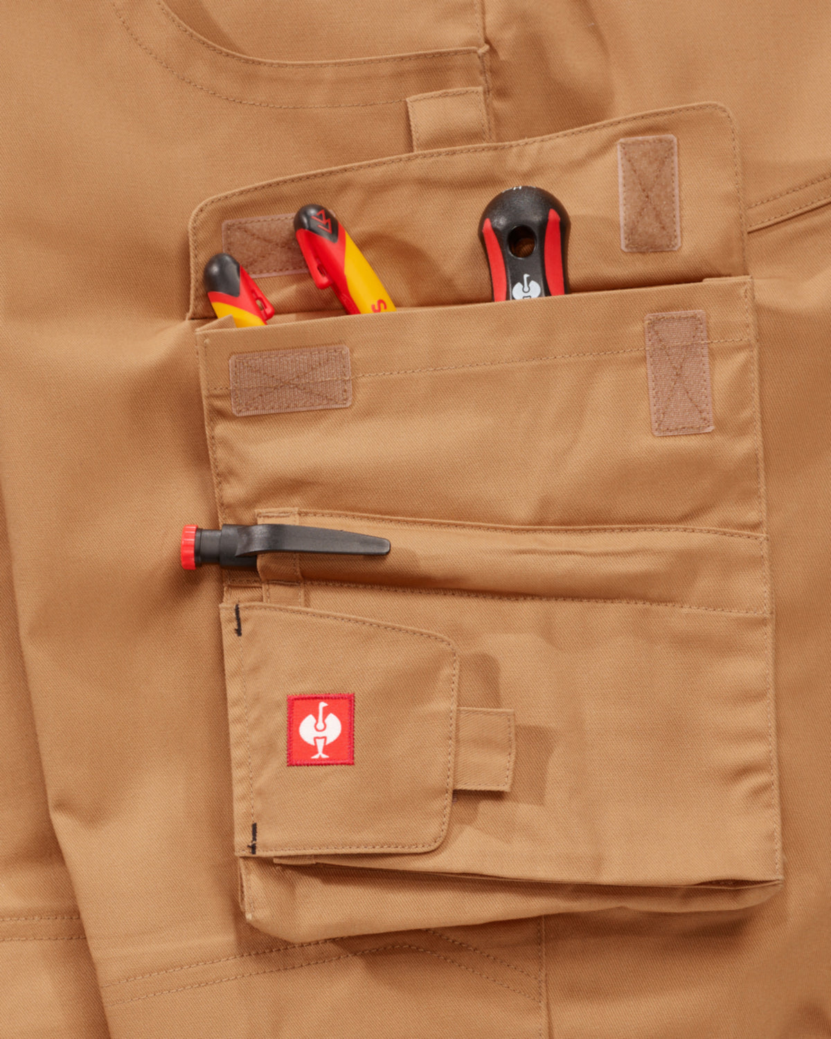 Close-up of a tan utility pocket on a matching tan garment, holding tools and a pen, with a small red square logo patch on the pocket flap.