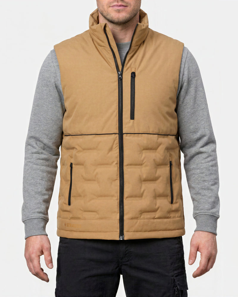 A man wearing a tan insulated zip-front vest over a gray long-sleeve shirt and black pants, shown from the lower face to upper thighs against a light gray studio background.