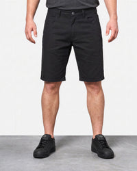 A model from the waist down wearing black shorts, a dark gray t-shirt, and black sneakers, standing against a white wall and gray floor.