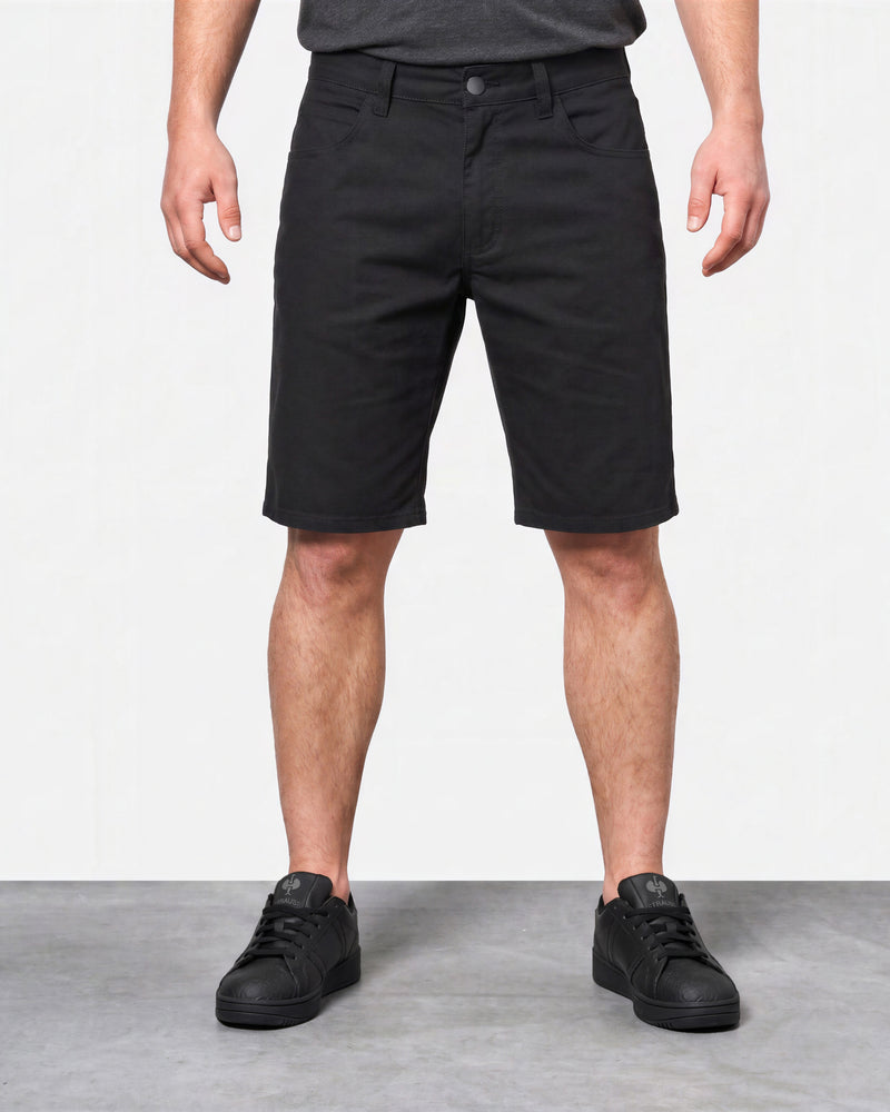 A model from the waist down wearing black shorts, a dark gray t-shirt, and black sneakers, standing against a white wall and gray floor.