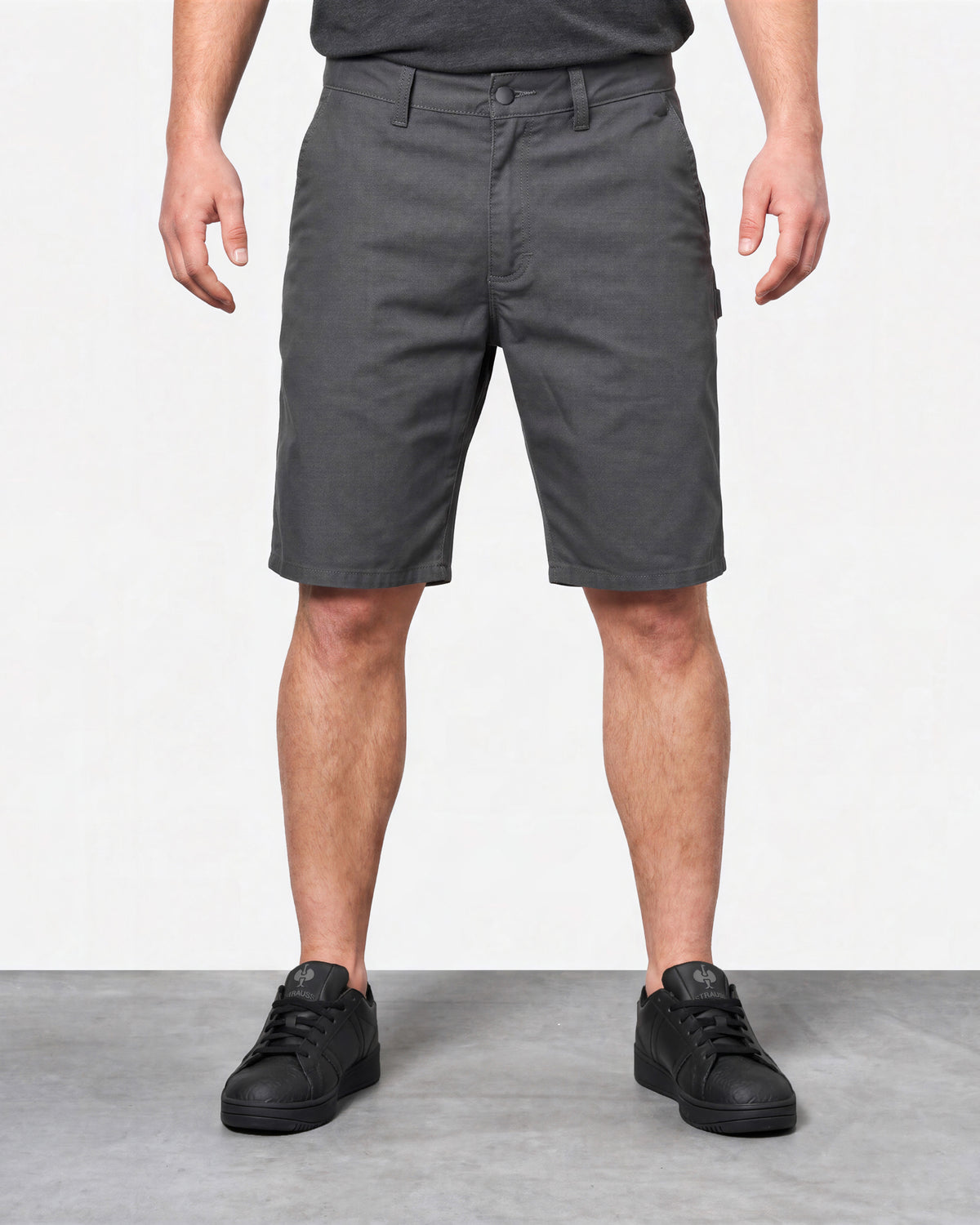 A person wearing dark gray knee-length shorts, a dark gray t-shirt, and black sneakers, standing against a light gray studio background.