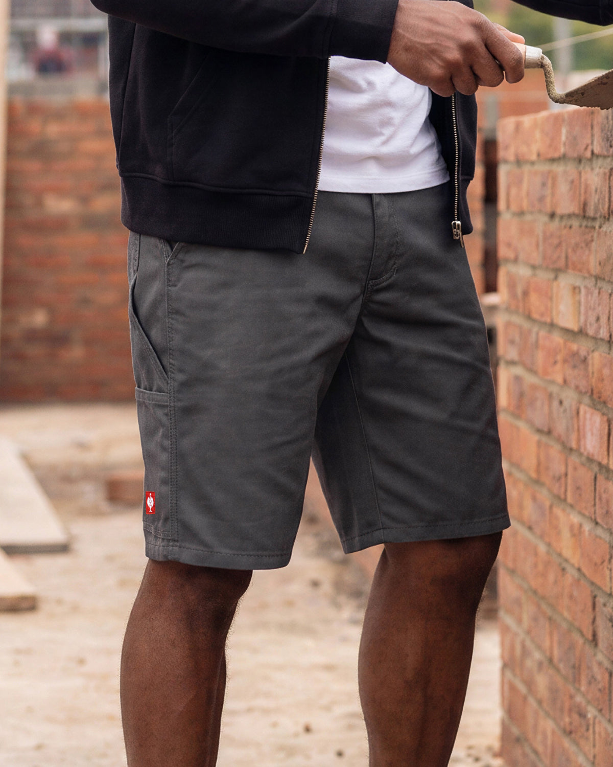 A person wearing dark gray cargo shorts, a white t-shirt, and a black zip-up jacket, standing next to a brick wall while holding a trowel.