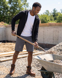 A man wearing a black zip-up hoodie, white t-shirt, gray shorts, and brown work boots smiles while holding a shovel next to a wheelbarrow at a construction site.